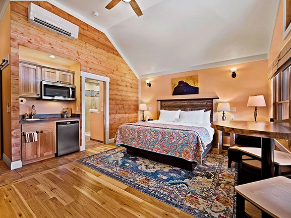 A wide view of a bungalow room with a bed, kitchenette, desk, and rug.