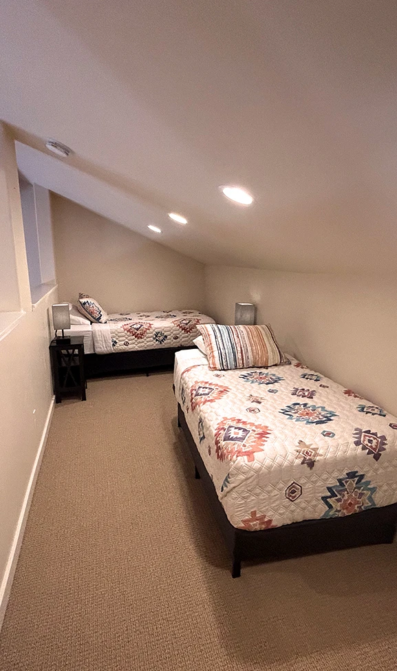 A small guest room with two beds, a nightstand, and a lamp.